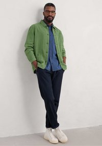 Green linen shirt jacket over a blue button-up shirt, paired with navy pants and white high-top sneakers. Distinct button details and texture.