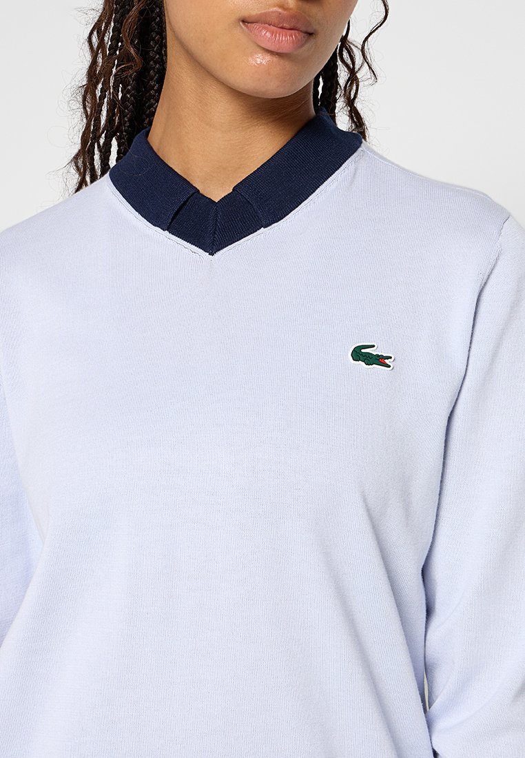 V-Ausschnitt-Pullover aus hellblauem Baumwollmaterial, mit einem marineblauen Kragen und einem kleinen grünen gestickten Krokodil-Logo auf der Brust.