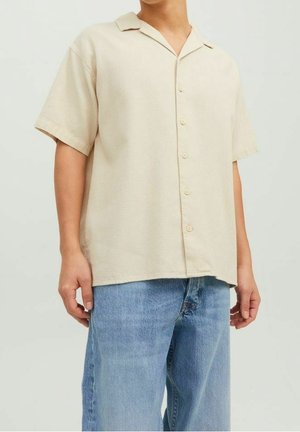 Osoba ubrana w beżową koszulę z krótkim rękawem zapinaną na guziki i jasnoniebieskie dżinsy, stojąca na prostym jasnym tle.