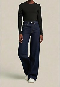 Personne portant un haut noir à manches longues, un jean bleu foncé taille haute à jambes larges et des chaussures blanches sur un fond beige uni.