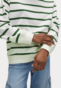 Grön- och vitrandig sweatshirt med ribbade ärmslut. Mjuk bomullsmaterial, avslappnad passform, synlig sömnad, bärs över ljusblå jeans.