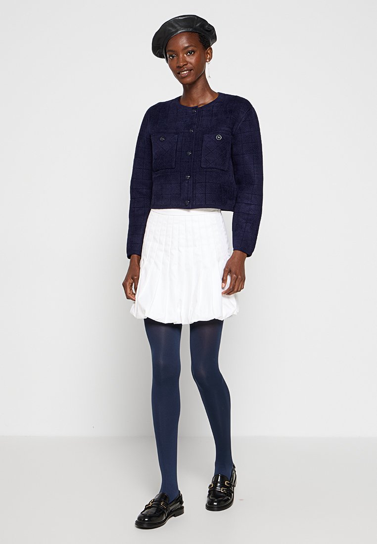 Granatowa, fakturowana kurtka o kroju cropped z dwoma kieszeniami na piersi, zestawiona z białą spódnicą plisowaną i granatowymi rajstopami, uzupełniona czarnymi butami i beretką.