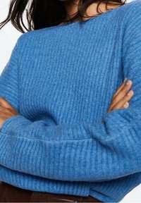 Frau mit verschränkten Armen, die einen blauen, gerippten Pullover mit langen Ärmeln und braunen Hosen trägt.