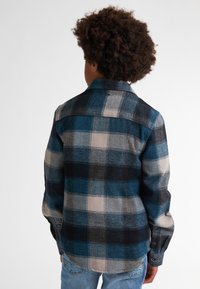 Camisa de franela a cuadros en tonos azul, negro y beige. Cuenta con un collar, puños con botones y un dobladillo recto. Textura suave y corte relajado.