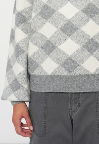 Grau- und cremefarbener kariert gestrickter Pullover mit geripptem Saum und weiten Ärmeln. Kombiniert mit grauen Cargo-Hosen, die über ein Detail mit Taschen verfügen.