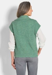 Grüner gerippter Strickpullover mit Schalkragen, kurzen Ärmeln und einer weichen Textur, über einem weißen Langarmshirt und grauen Jeans getragen.
