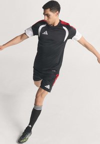 Young male soccer player wearing black Adidas kit and cleats, mid-kick with arms extended against a plain background.