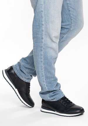 Beine, die hellblaue Jeans und schwarze Schnür-Sneaker mit weißen Sohlen tragen, ein Fuß leicht angehoben vor weißem Hintergrund.
