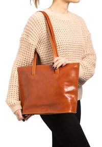 Bruine leren tote bag met een gladde textuur en een gestructureerde vorm. Voorzien van twee handvatten en zichtbare stiksels langs de randen.