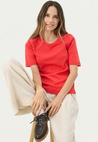 T-shirt rouge à manches courtes au design ajusté, fabriqué en tissu lisse. Associé à un pantalon large beige clair et des chaussures à lacets marron.