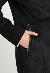 Cappotto in suede nero con tasca laterale con zip e un morbido rivestimento interno visibile all'orlo delle maniche. Texture liscia e design su misura.