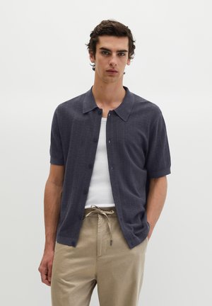 Jonge man met een marineblauw kort mouw knoophemd over een wit hemdje en beige broek met trekkoord, staand tegen een effen achtergrond.