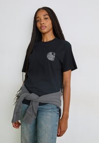 Black cotton t-shirt with short sleeves, featuring a gray graphic design on the left chest. Paired with loosely tied gray sweatshirt around the waist.