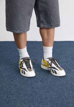 White and gray sneakers with yellow accents, textured overlays, and gray laces. Worn with gray shorts and white ankle socks on blue carpet.