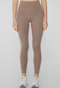 Leggings marrones, ajustados y elásticos, con una cintura alta y textura suave. Combinados con zapatillas deportivas blancas y grises.