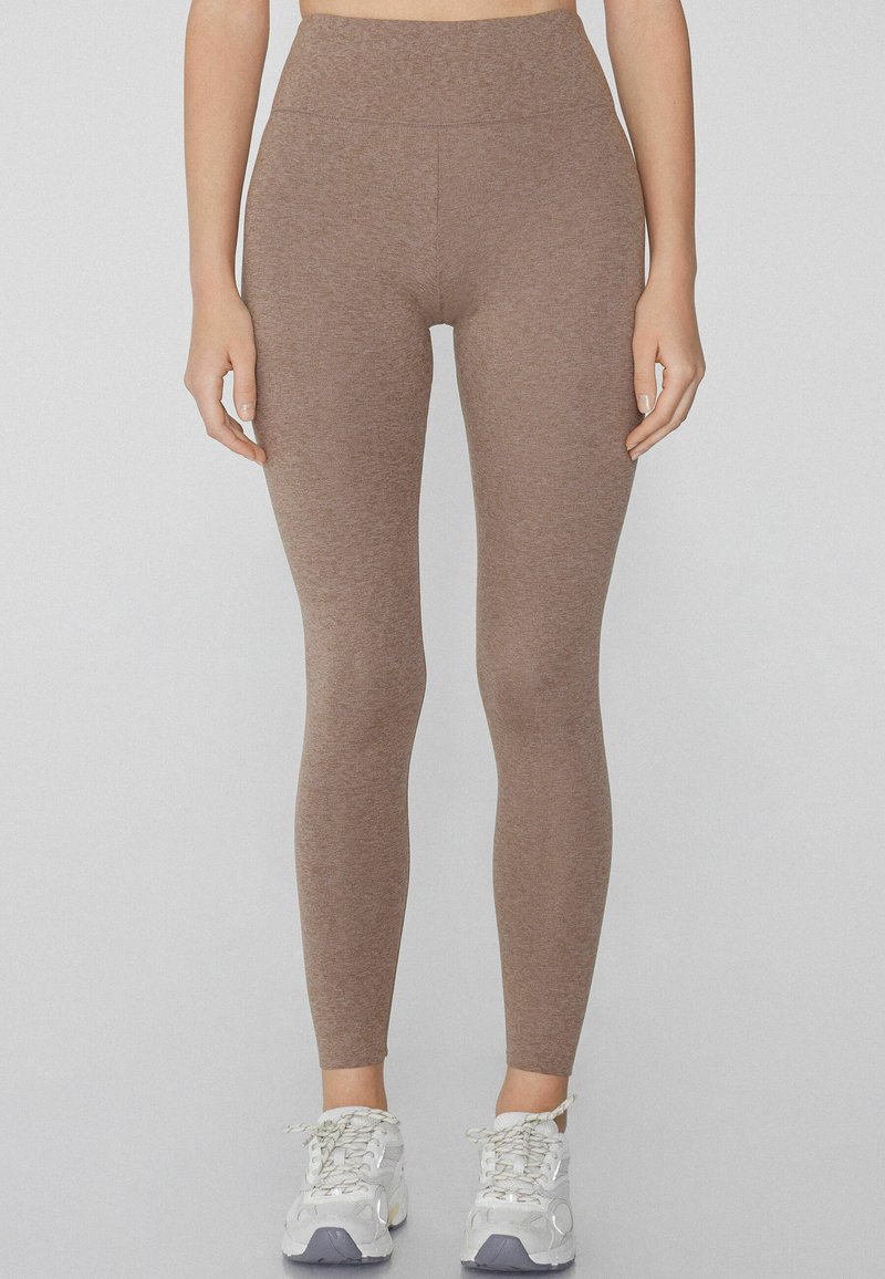Leggings marrones, ajustados y elásticos, con una cintura alta y textura suave. Combinados con zapatillas deportivas blancas y grises.