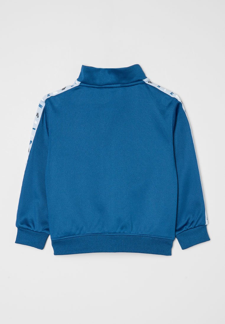 Chaqueta de pista azul con un cuello alto, puños elásticos y franjas laterales blancas que presentan pequeños logotipos negros. Tejido suave y ligero.