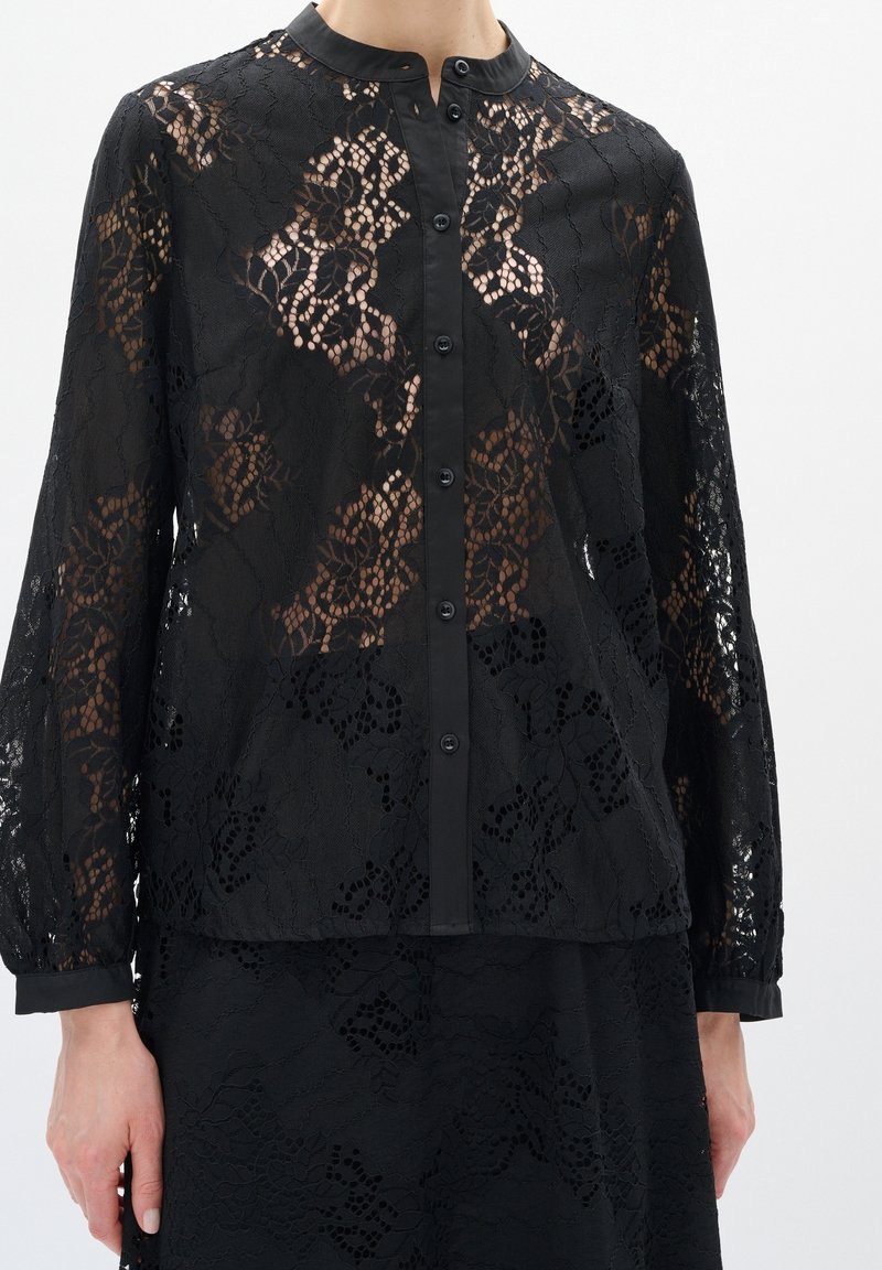 Blouse noire en dentelle à manches longues avec motif floral, devant boutonné et col sans col, portée avec une jupe en dentelle assortie.