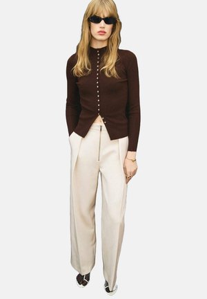 Cardigan lungo a maniche raglan marrone a coste con dettagli in bottone, abbinato a pantaloni a gamba larga crema e occhiali da sole neri.