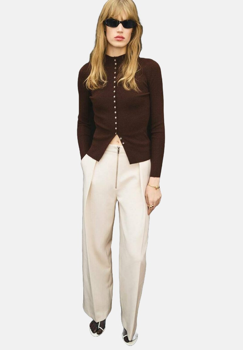 Cardigan lungo a maniche raglan marrone a coste con dettagli in bottone, abbinato a pantaloni a gamba larga crema e occhiali da sole neri.