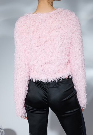 Osoba ubrana w puszysty różowy sweter z długim rękawem i błyszczące czarne spodnie, widziana od tyłu na neutralnym tle.