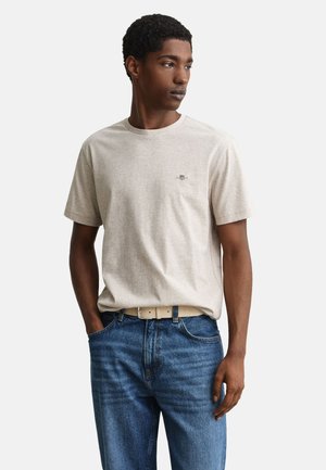 Giovane uomo che indossa una maglietta beige a maniche corte con un piccolo logo sul petto, jeans blu e cintura beige, in piedi con una mano in tasca.