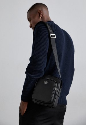 Sort læder crossbody taske med en lynlåslomme, logodetalje og en justerbar rem med brandnavn. Båret over en tekstureret marineblå sweater.