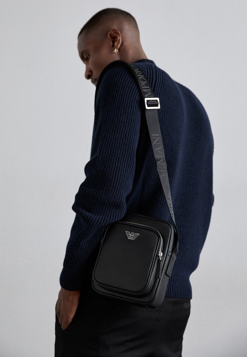 Svart läder crossbody-väska med ett zippered fack, logodetalj och en justerbar axelrem med märke. Bärs mot en strukturerad marinblå tröja.