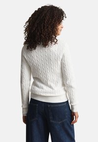 Biely sveter s vrkočovým vzorom, s rebrovaným lemom a manžetami, zdobený textúrovaným vzorom. Nosený s tmavomodrými širokými riflemi.