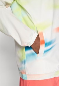 Lättviktsvit jacka med multicolor gradientmönster, med elastiska ärmslut och sidofickor. Mjuk textur.