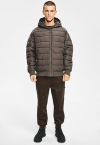 Brązowa pikowana kurtka z kapturem, z zamkiem błyskawicznym z przodu i ściągaczami na mankietach, w połączeniu z ciemnobrązowymi dresami i czarnymi butami.