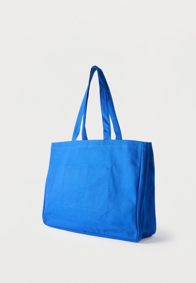 Μπλε τσάντα tote από ύφασμα με δύο λουριά ώμου και ένα ραμμένο ορθογώνιο έμπλαστρο στο μπροστινό μέρος, παρουσιάζεται πάνω σε ένα απλό ανοιχτό χρώμα φόντο.