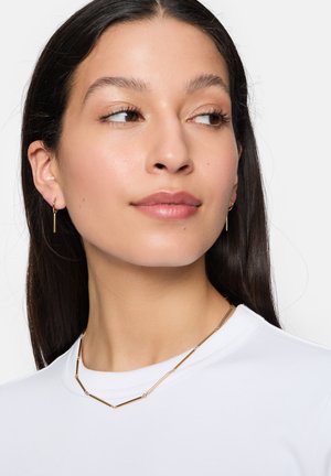 Goldfarbene Schmuckstücke mit minimalistischem Design. Dünner, rechteckiger Anhänger an einer zarten Kette, getragen zu einem weißen Hemd.