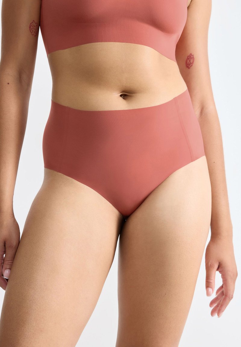 Kvinna som bär högmidjade, sömlösa underkläder i rostfärg och matchande bralette, visar midja och övre lår, med små arm-tatueringar.