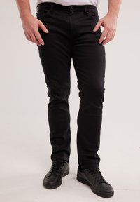 Férfi fekete testhezálló nadrágban és fekete fűzős cipőben, világos háttér előtt áll.