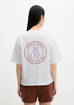Femme aux longs cheveux noirs porte un t-shirt blanc oversize avec un logo circulaire "FC Marc O'Polo Stockholm 1967" et un short marron.