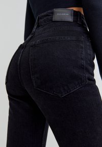 Jean en denim noir taille haute avec une texture lisse, un design classique à cinq poches et un écusson en cuir marqué à la ceinture.
