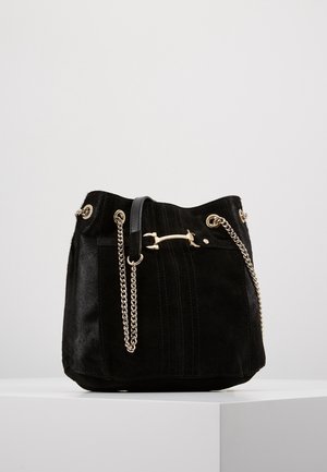 Sac seau en daim noir avec une bandoulière chaîne dorée et un fermoir barre doré, présenté sur une surface blanche devant un fond gris clair.