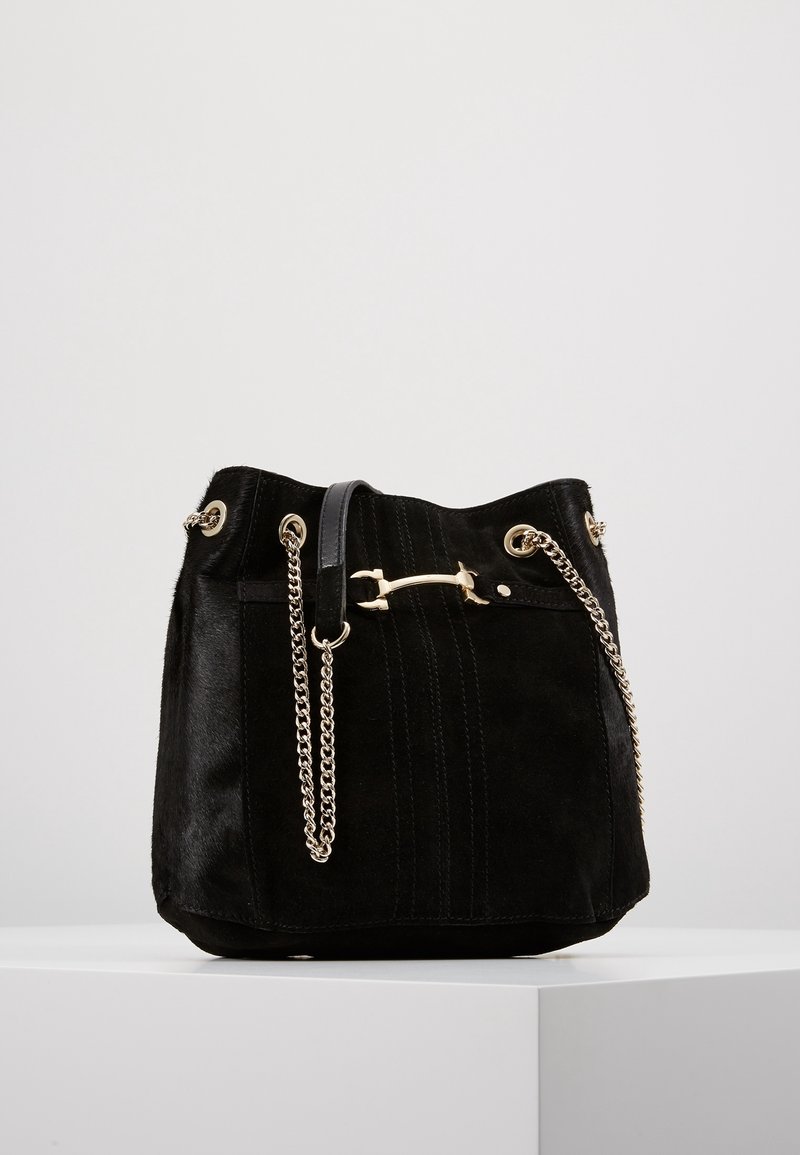 Sac seau en daim noir avec une bandoulière chaîne dorée et un fermoir barre doré, présenté sur une surface blanche devant un fond gris clair.