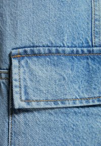 Ljusblå denimtyg med synlig textur, med en fyrkantig klaffficka och kontrasterande orange sömmar längs kanterna och sömmarna.