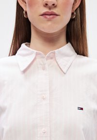 Svijetlopinkasta košulja s kragnom, s vertikalnim bijelim prugama, gumbima i malim vezom u mornarskoj plavoj, crvenoj i bijeloj boji na prsima.