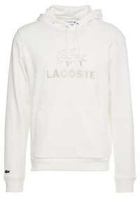 Vit hoodie av mjukt tyg med en stor "LACOSTE"-logotyp och en krokodilgrafik i krämfärg, känguruficka och huva med dragsko.