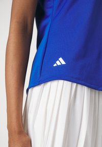 Modra športna majica iz gladkega materiala z belim logotipom Adidas na robu. Utkana z belo nabranimi krilom.