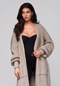 Langer, offen gestrickter Cardigan in hellbeige, mit strukturiertem Stoff, oversized Ärmeln und vorderen Taschen, kombiniert mit einem schwarzen Body.