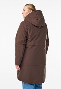 Femme de profil à gauche portant un manteau d'hiver à capuche marron avec des manches longues et un jean bleu sur fond blanc.