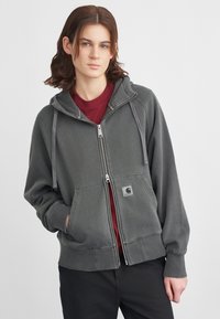 Sudadera gris con cremallera hecha de un material suave, con un bolsillo frontal, cordones en la capucha y un parche con el logo. Llevada sobre una camiseta color burdeos.