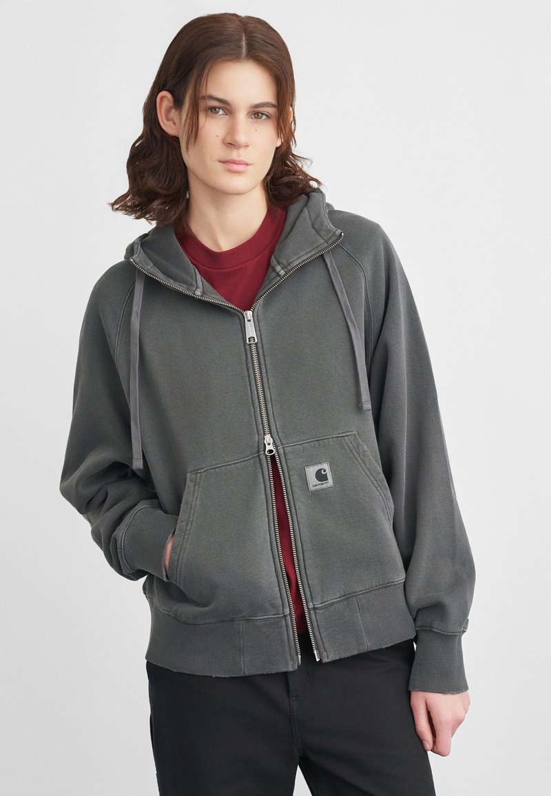 Sudadera gris con cremallera hecha de un material suave, con un bolsillo frontal, cordones en la capucha y un parche con el logo. Llevada sobre una camiseta color burdeos.