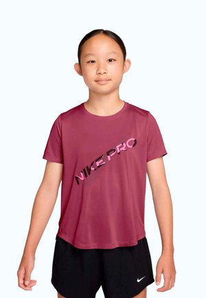 Junge Person mit dunklen, zurückgebundenen Haaren, die ein dunkelrotes Nike Pro T-Shirt und schwarze Nike Shorts trägt und vor einem schlichten Hintergrund steht.