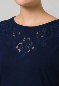 Žena v námořnickém modrém topu s vyšívanými květinovými krajkovými detaily u výstřihu na světlém pozadí.