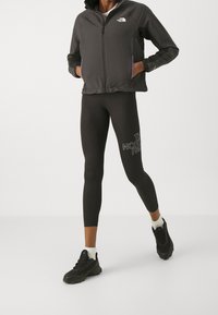 Person som bär en svart zip-up jacka och svarta leggings med The North Face-loggan, i kombination med svarta sneakers och vita strumpor.
